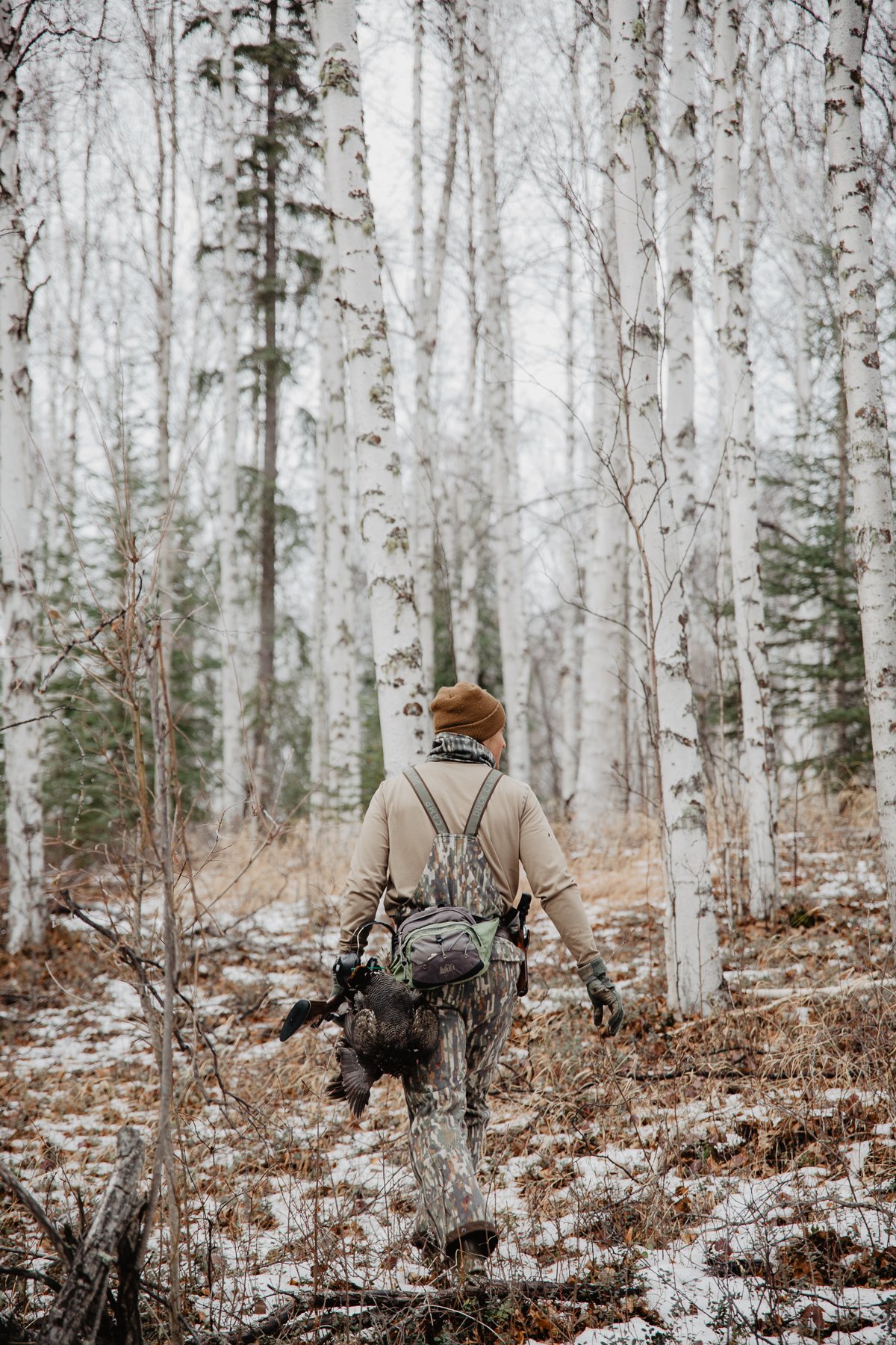 Hunter walks away from viewer in winter forest with dead bird in hand. 