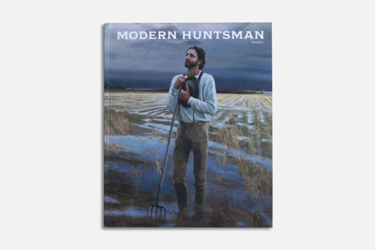 Aerial shot of a man standing in a flooded rice field holding a pitchfork during overcast weather, featured on Modern Huntsman's magazine cover emphasizing sustainable hunting, nature, and outdoor lifestyle.